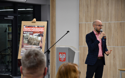 człowiek mówiący do mikrofonu stojący obok mównicy z godłem Polski i tablicy z plakatem "Front Północny Powstania Wielkopolskiego"